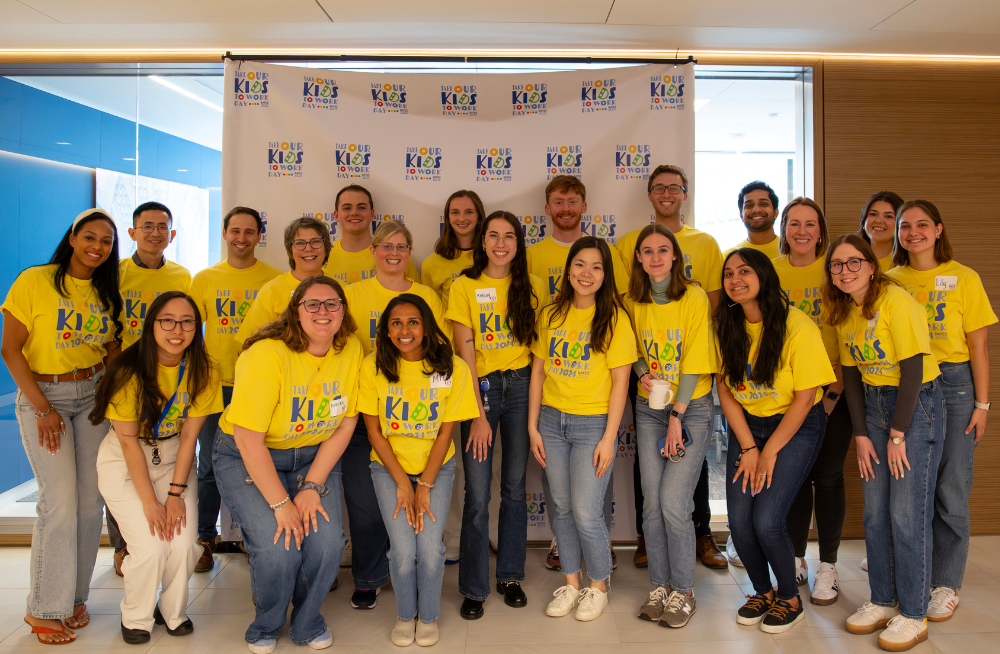 A large group of men and women standing together and smiling at the camera. A white backdrop behind them, as well as their shirts, have the label: "Take Our Kids to Work Day."