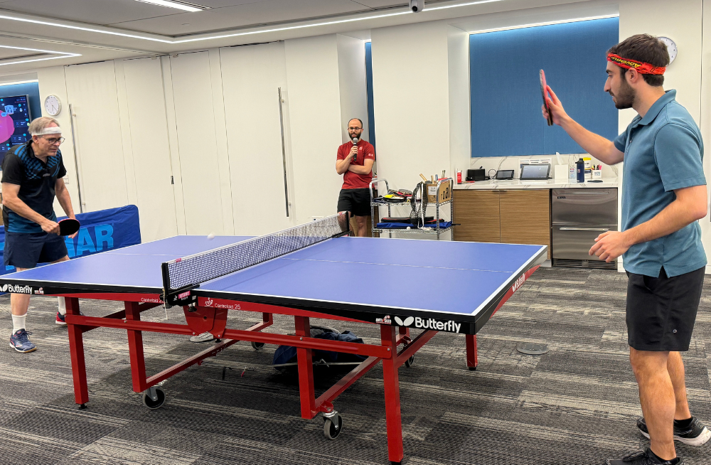 An action shot of two men playing ping-pong. A referee stands in the background watching with a microphone.