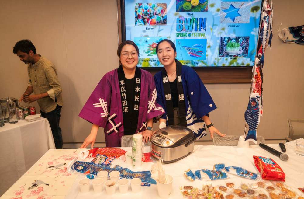 Two women in robes with East Asian symbols. They are smiling and standing behind a table clad with food and a rice cooker.