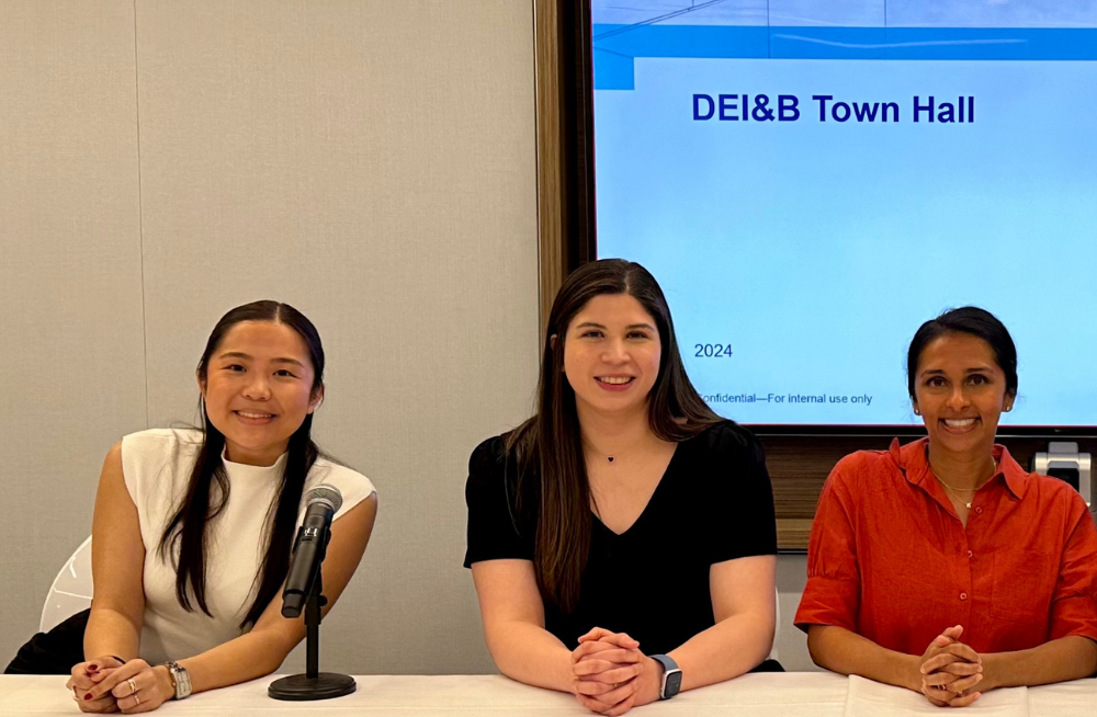 Three women seated behind a table, smiling at the camera. The table has a microphone on it, and behind the table is a screen reading, "DEI&B Town Hall."