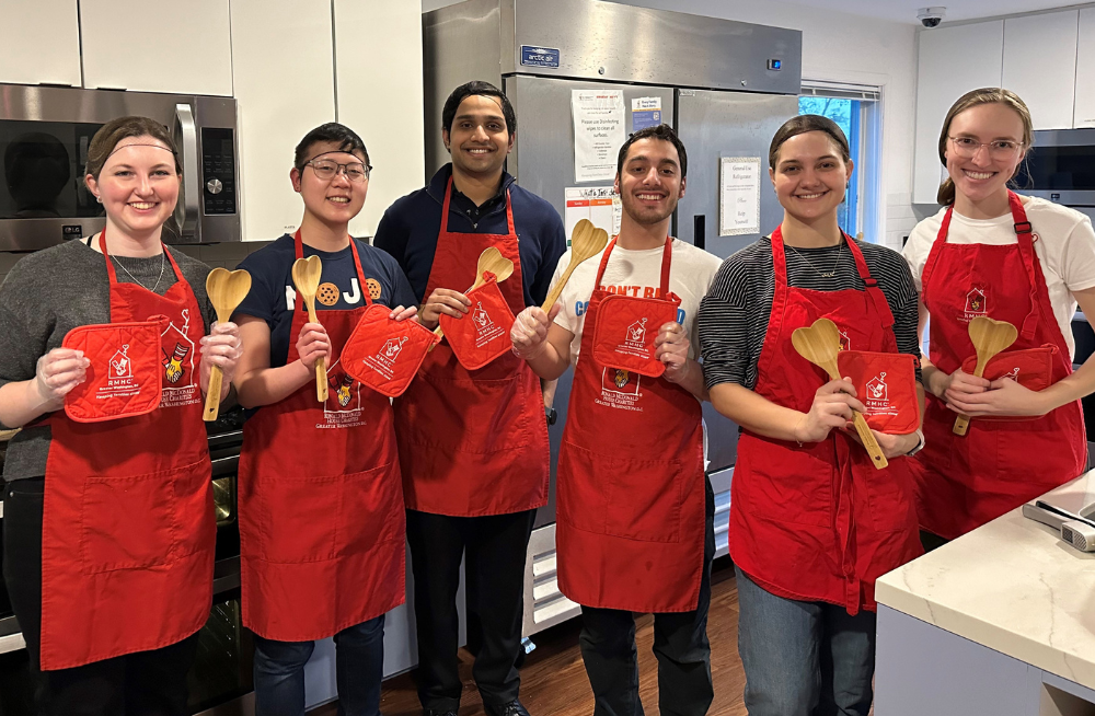 A group of young men and women wearing red aprons clad with the Ronald McDonald House logo. They are holding over mitts and heart-shaped spoons. They are standing and smiling in a large kitchen.