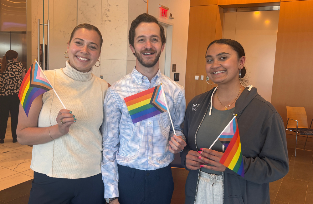Two young women and one young man standing together and smiling at the camera. They are holding rainbow-colored Pride flags.