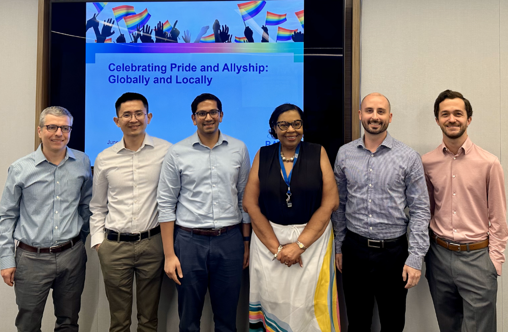 5 men and one woman stand together, smiling. The screen behind them reads: "Celebrating Pride and Allyship: Globally and Locally."