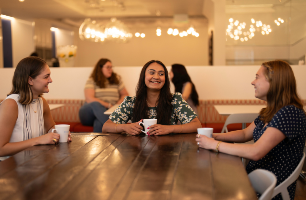 Three smiling women sitting around a table and holding coffee mugs.