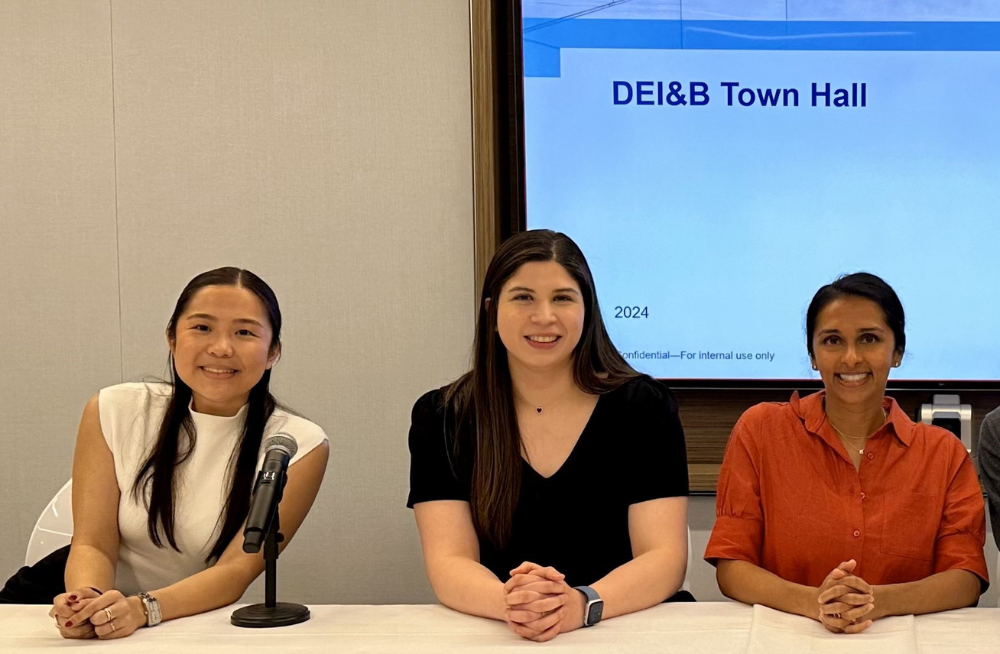 Three smiling young women sit behind a table, topped with a microphone. The screen behind them reads: "DEI&B Town Hall."