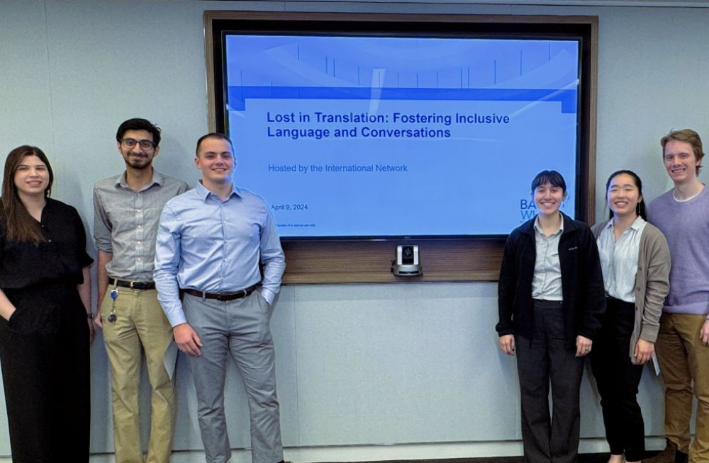 A group of three men and three women stand around a screen, smiling at the camera. The screen reads: "Lost in Translation: Fostering Inclusive Language and Conversations."