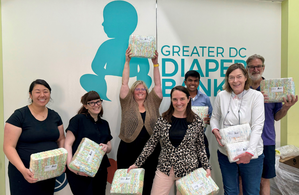 A group of men and women stand together smiling and holding packs of diapers. Signage on the wall behind them reads: "Greater DC Diaper Bank."
