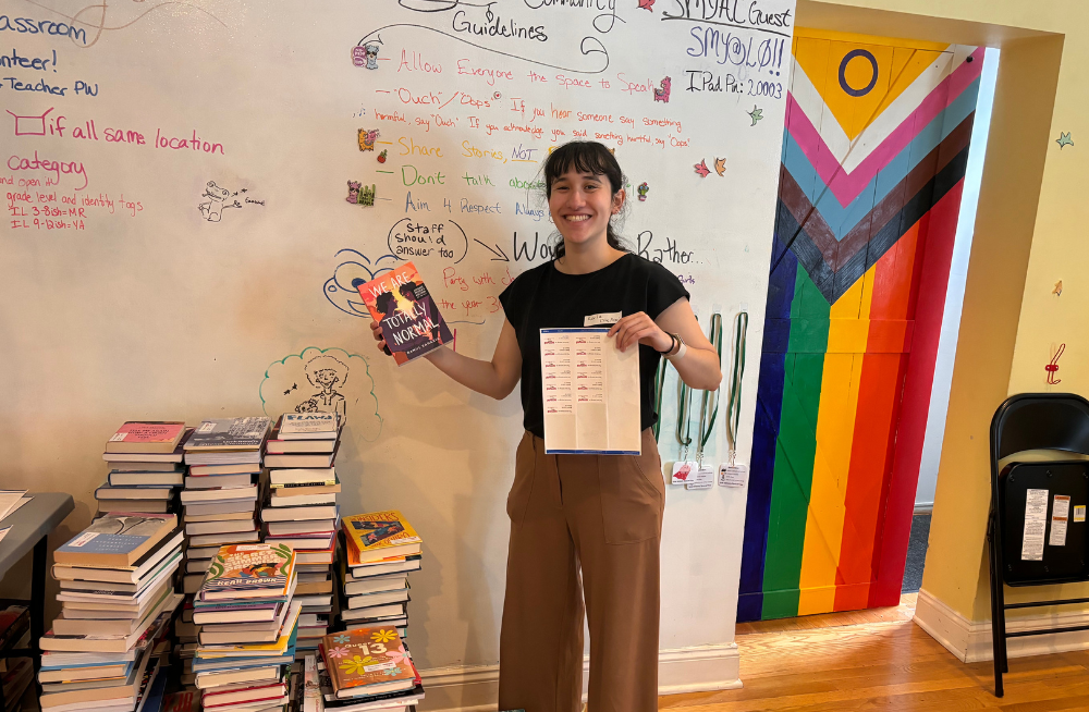 A young smiling woman stands next to a large pile of books. She is holding book labels in one hand and a book titled "We Are Totally Normal" in the other.