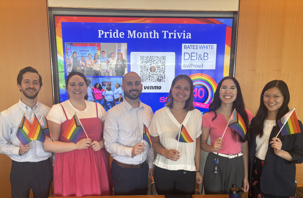 A group of young men and women smiling and standing together. They all are holding rainbow-colored pride flags, and behind them, a screen reads: "Pride Month Trivia."