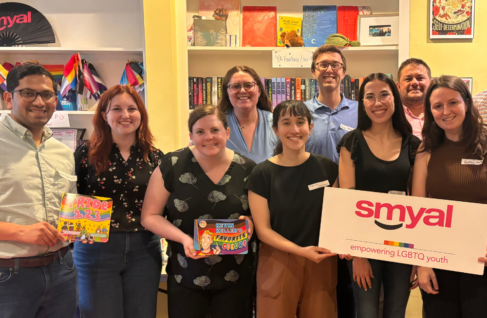 A group of men and women standing together and smiling at the camera. A bookshelf is behind them. Some of the people are holding books; others are holding a sign labeled "smyal."