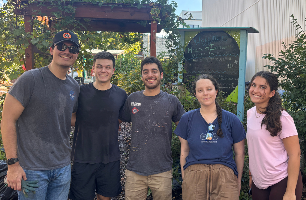 Three young men and two young women stand together smiling at the camera. They are outside; bushes and other foliage are behind them.