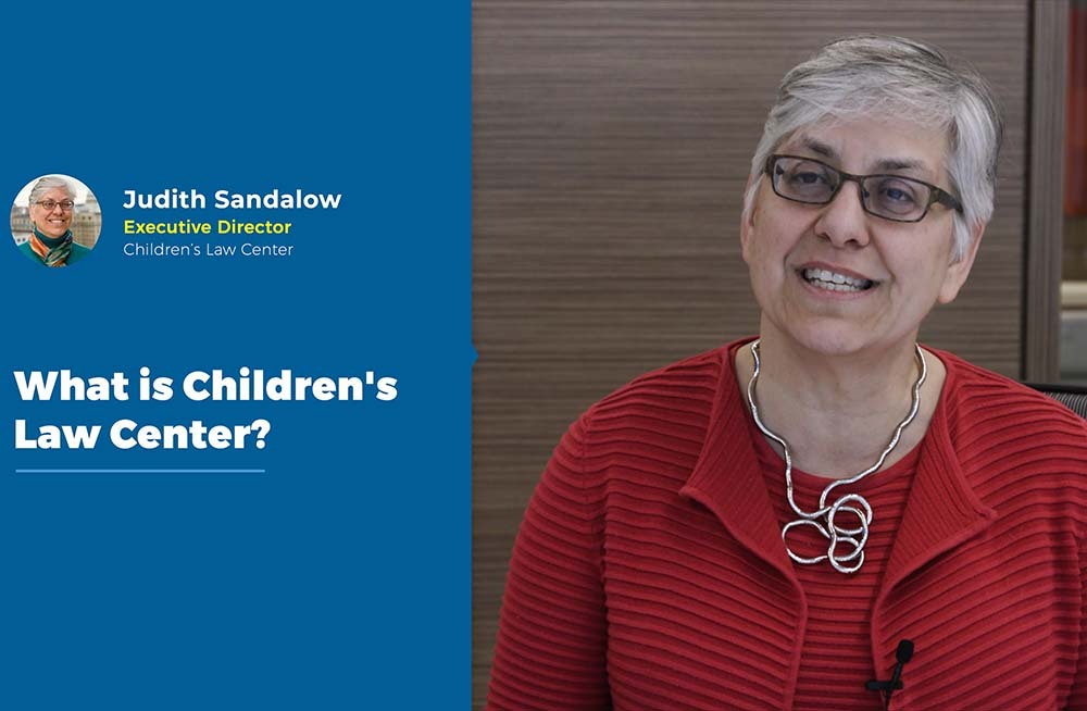 woman in red dress and glasses speaking; sign on left of image says What is Children's Law Center