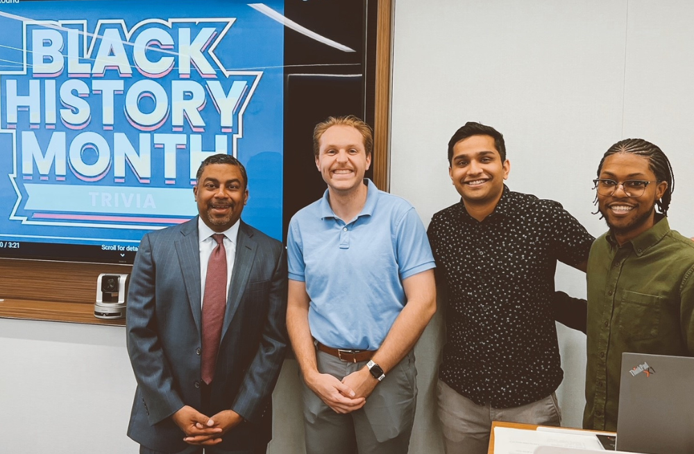 Four men stand together, smiling at the camera. A screen behind them reads: "Black History Month Trivia."