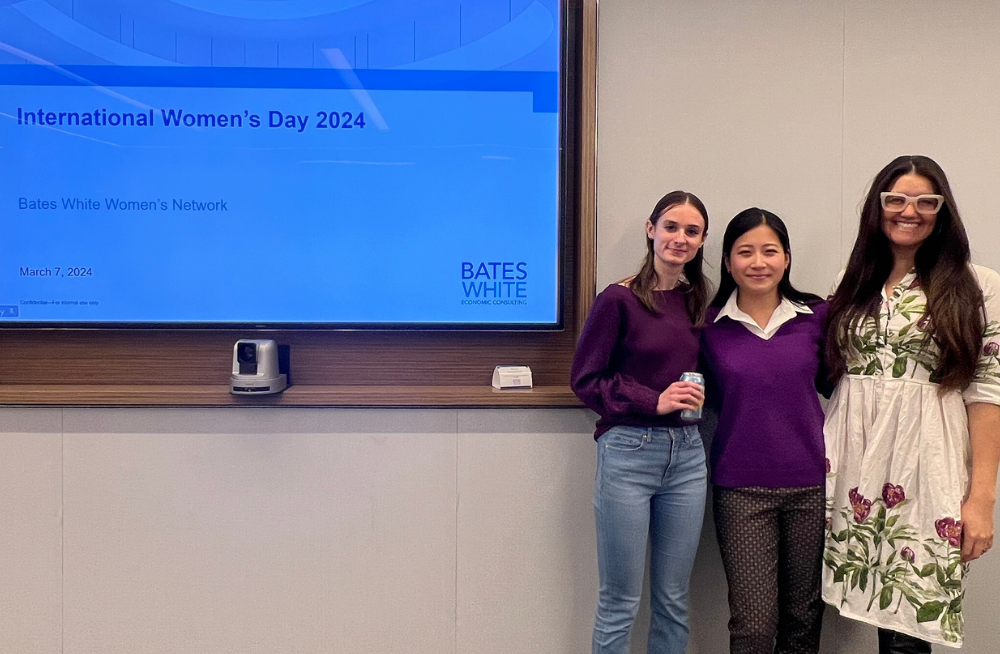 Three young women standing together and smiling. The screen next to them is titled, “International Women’s Day 2024.”