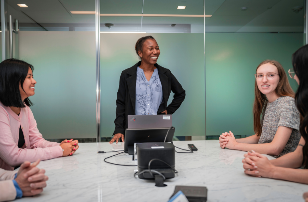 A group of smiling women sit around a large table. One woman stands behind a computer at the head of the table.