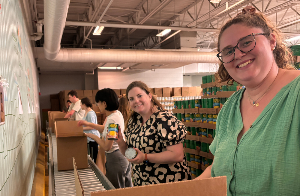 Several young men and women are lined up around a conveyor belt clad with boxes. They are putting cans into these boxes. Two of the women are smiling at the camera.