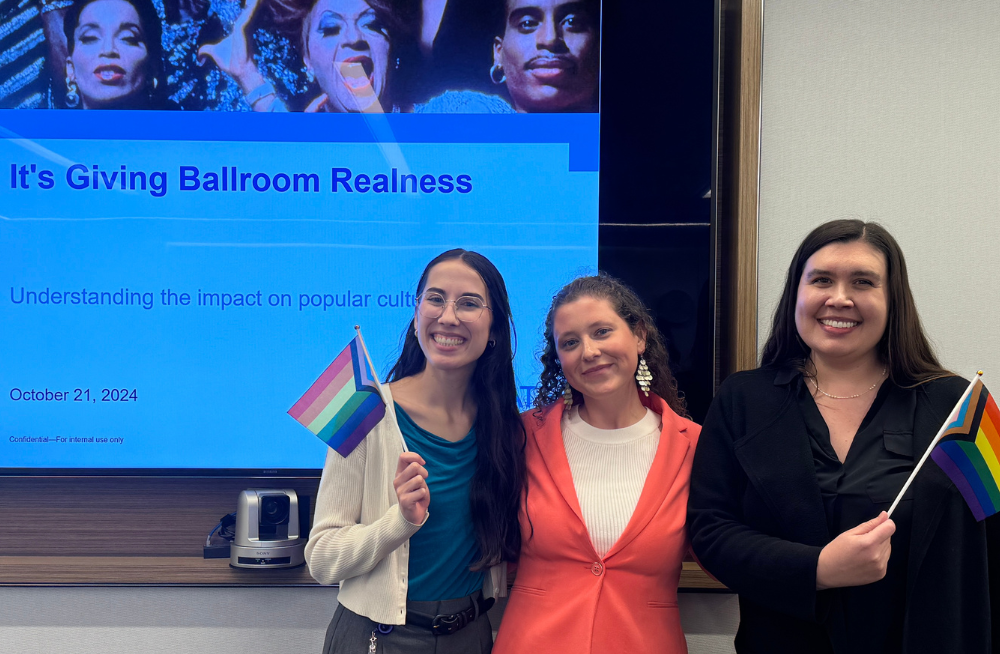 Three women stand smiling at the camera; two of them hold pride flags.  A screen behind them is titled "It's Giving Ballroom Realness."