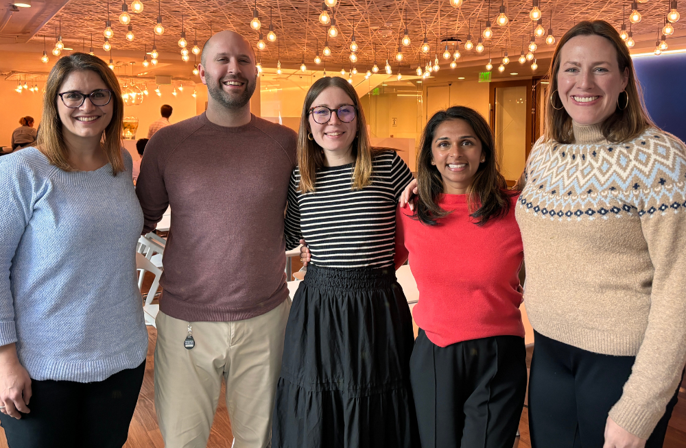 Four women and one man stand arm-in-arm together, smiling at the camera.