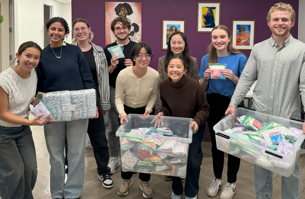 A group of 9 young men and women stand together, smiling at the camera. Many of them are holding large boxes or individual bags of hygiene products.