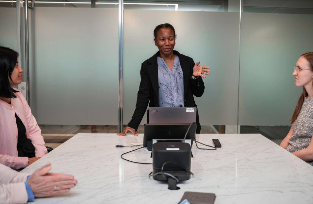 A young woman stands at the end of a long table, in the middle of a conversation. Women on either side of the table are seated, listening to her.