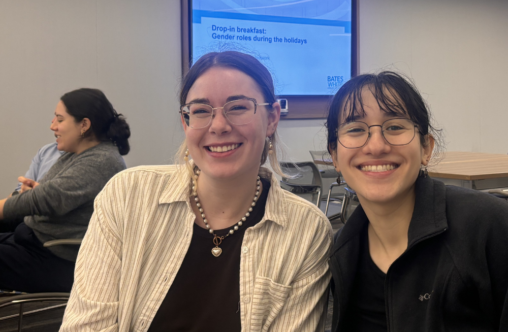 Two young women are sitting next to each other and smiling at the camera. A screen behind them reads, “Drop-in breakfast: Gender roles during the holidays.”