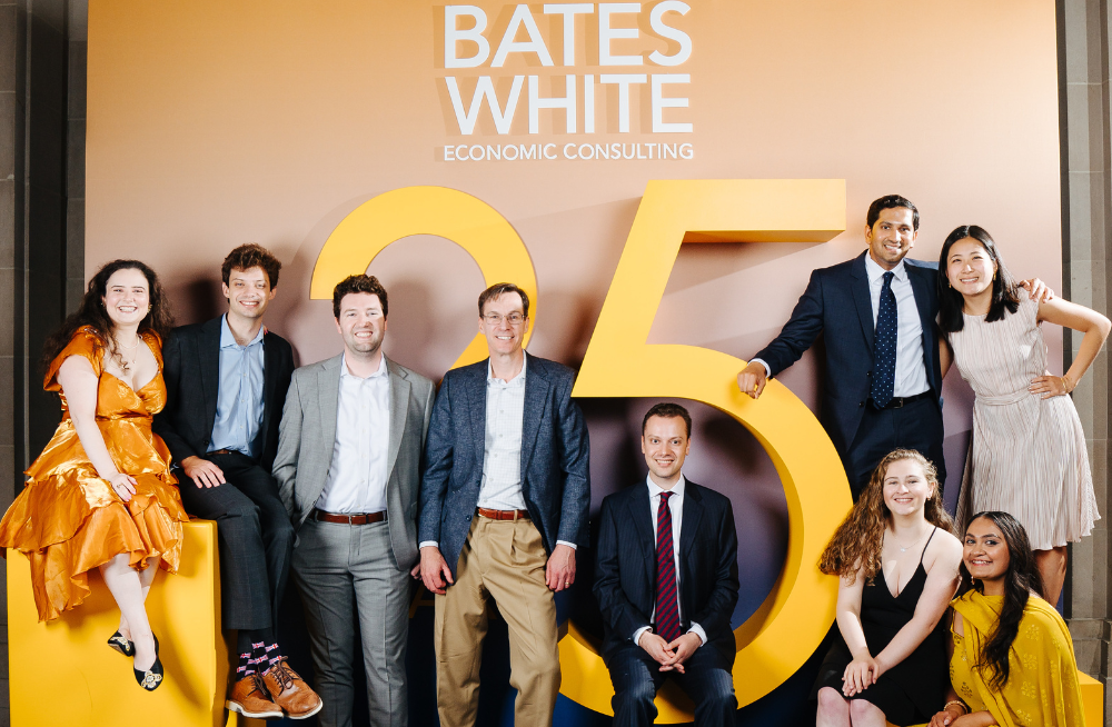 A group of men and women dressed in formal clothing are gathered together and smiling. A sign behind them reads: "Bates White Economic Consulting 25."