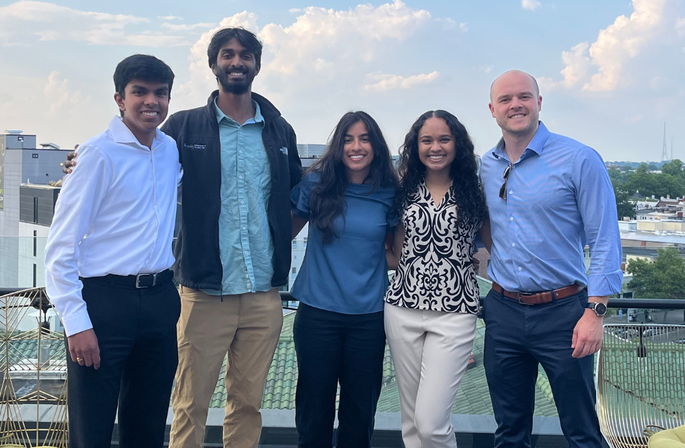 A group of young men and women are smiling at the camera, arm-in-arm. Behind them is a rooftop view of a city.