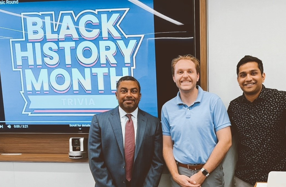 Three men stand together smiling at the camera. A screen behind them reads "Black History Month Trivia."