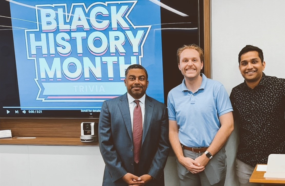 Three men stand together smiling at the camera. A screen behind them reads: Black History Month Trivia.