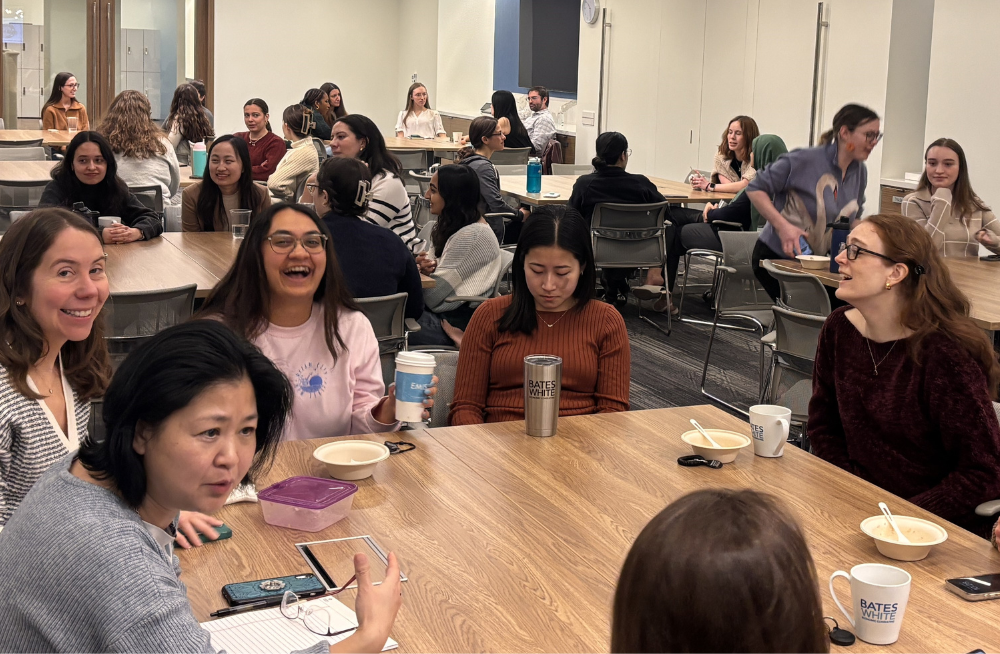 A group of women are gathered around the table and smiling. Behind them are more tables surrounded by people.