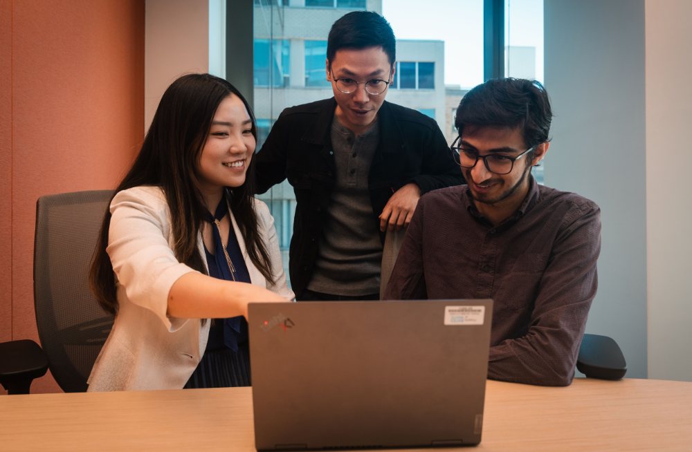 A young man and woman sitting together, smiling at a computer screen. A young man stands behind them, also looking at the screen.