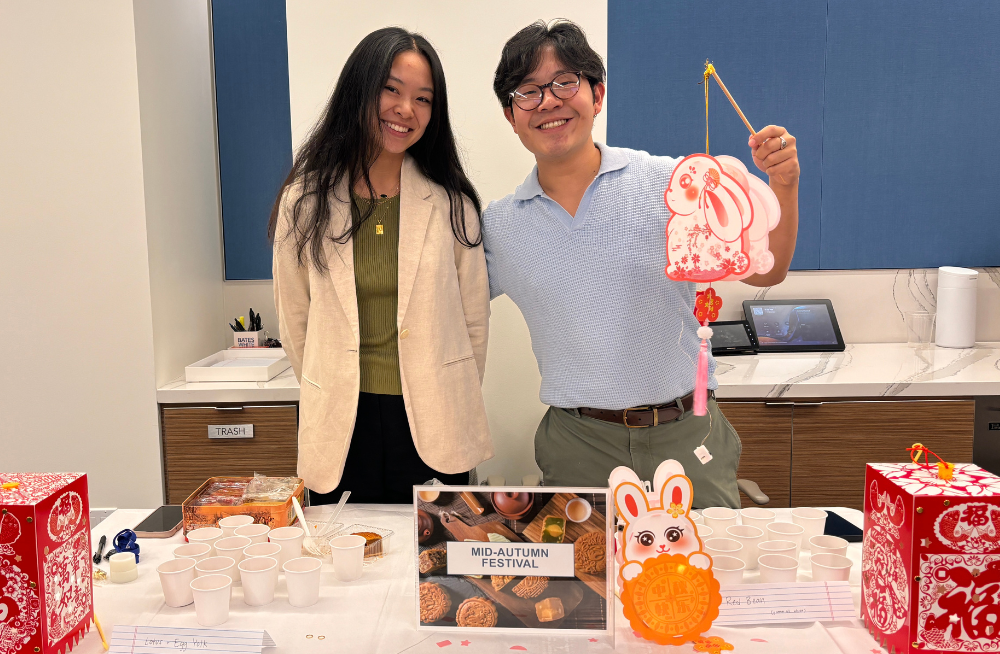 A young man and a young woman stand behind a table smiling at the camera. The table has cups of food and a sign reading: “Mid-Autumn Festival.” The man holds a paper cut-out of a bunny.