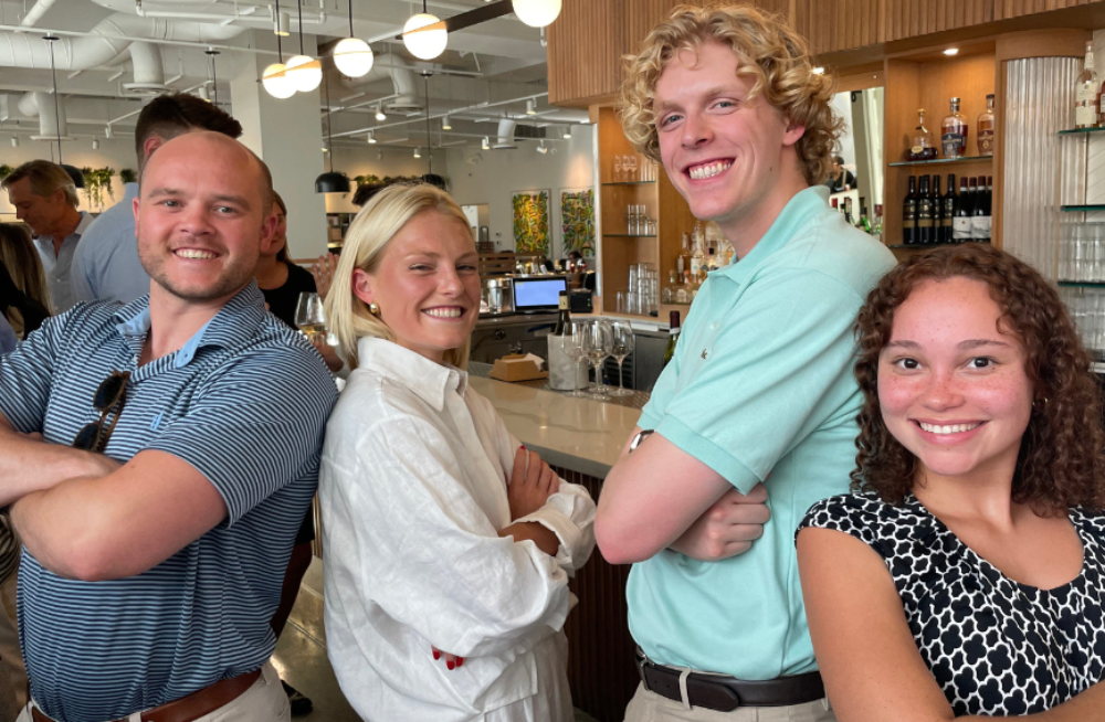 Two young men and two young women are standing together smiling at the camera with their arms crossed. The background shows a restaurant and other people.