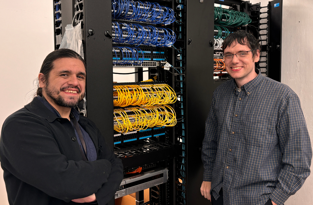 Two men stand together, smiling at the camera. Behind them is a shelf-like object containing wires and outlets.