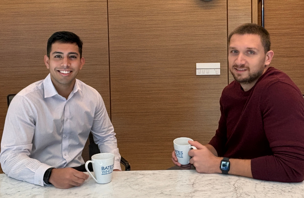 Two young men are seated behind a table, each with a coffee mug imprinted with the Bates White logo. Both are smiling at the camera.