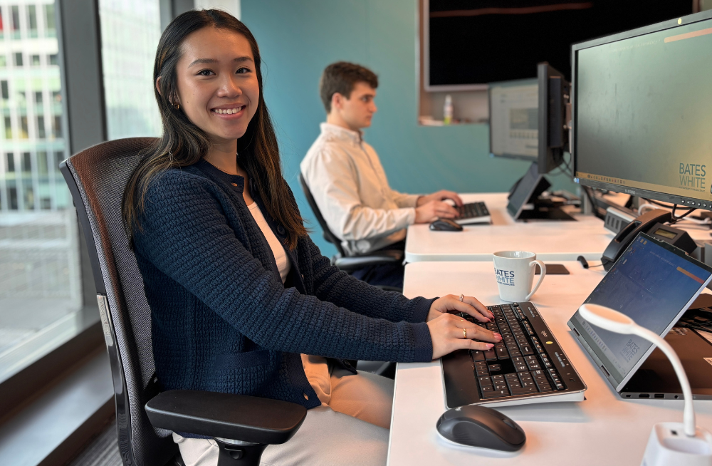 A young woman is seated in front of a laptop and monitor. In the middle of typing, she is turning at the camera and smiling. Behind her is a man who is also typing on his own separate monitor.