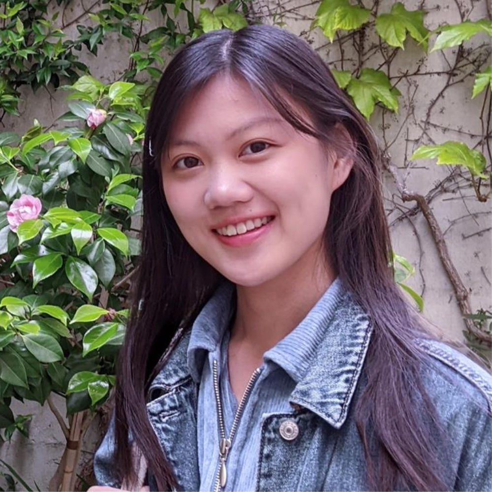 A smiling young woman looks at the camera. The background is a wall covered in foliage.