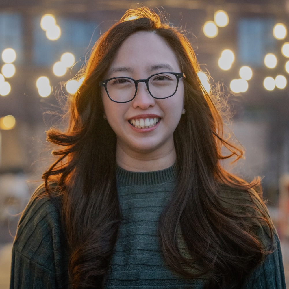 A young woman smiles at the camera. A blurred background behind her shows hanging lights and a building.