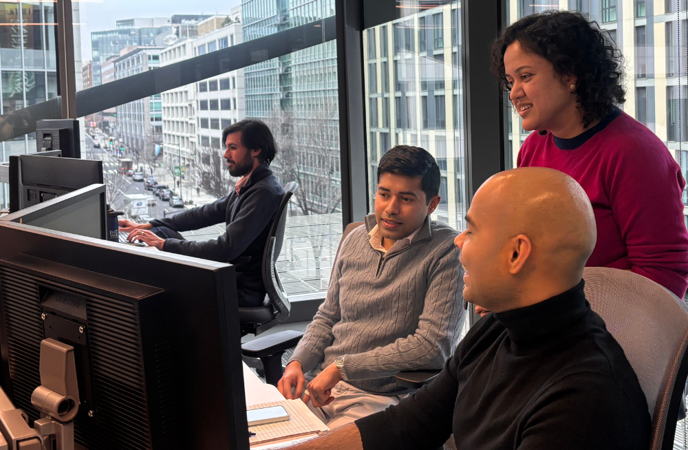 Two men are seated at a desk, staring at computer monitors. One man is in the middle of speaking to the other man and a woman standing behind them. Wide windows around them show an urban background.